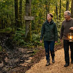 Zwei Menschen auf einem Waldweg, neben ihnen ist ein Schild "Der neue Weg beginnt hier.".